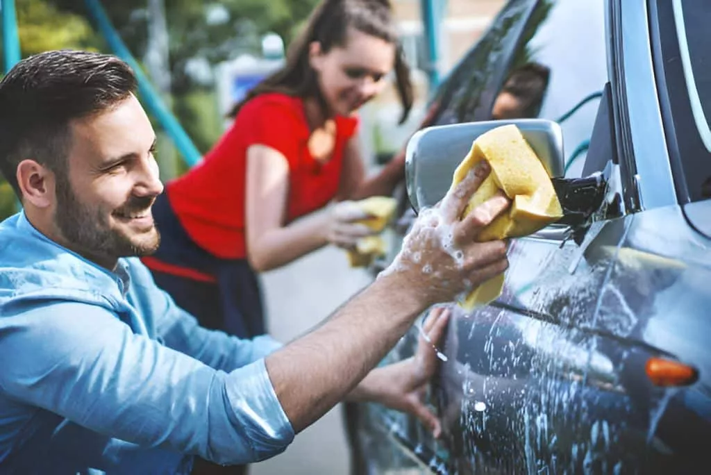 Couple washing a car together using yellow sponges, smiling and having fun.