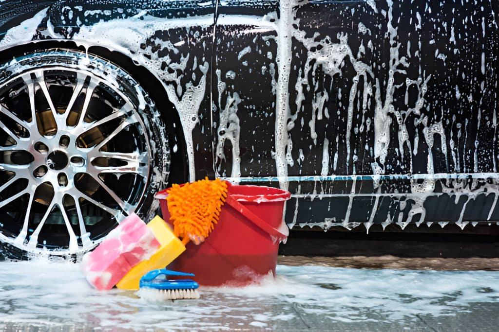 Car being washed with soap suds, showcasing a red bucket, chenille mitt, and other car cleaning tools.