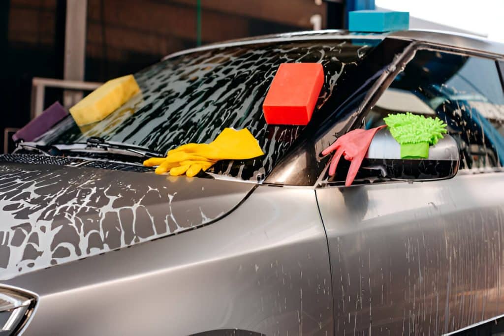 Various car cleaning tools including sponges, gloves, and mitts placed on a soapy car hood.